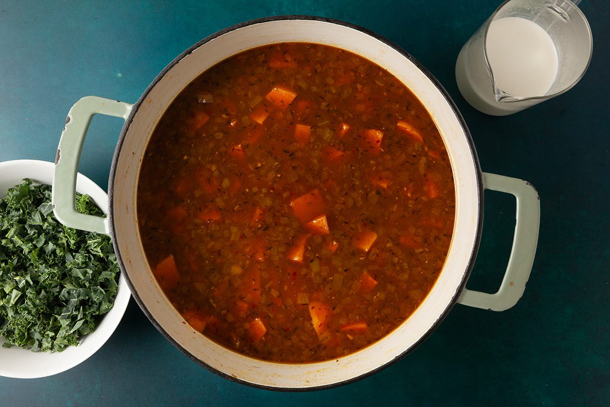 Overhead shot of a pot of vegetable soup with diced carrots on a teal surface, next to a bowl of chopped greens and a glass pitcher of milk or cream;