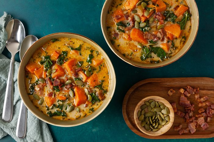Overhead shot of two bowls of Easy Sweet Potato Chowder filled with tender sweet potato chunks, leafy greens, crispy bacon, and pumpkin seeds in a creamy, golden broth; A wooden board with chopped bacon and a small bowl of pumpkin seeds sits beside the bowls on a dark teal surface;