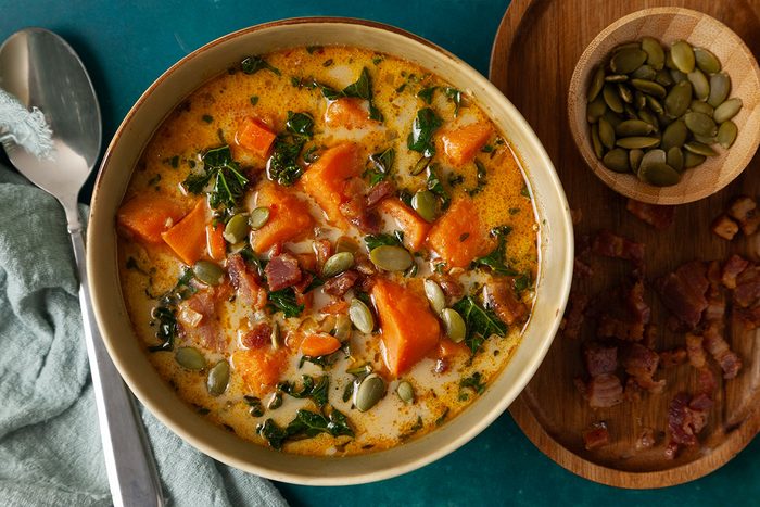 Overhead shot of Easy Sweet Potato Chowder, a creamy bowl filled with tender sweet potato chunks, wilted greens, crispy bacon pieces, and pumpkin seeds, set beside a wooden board holding extra bacon and a small bowl of pumpkin seeds;