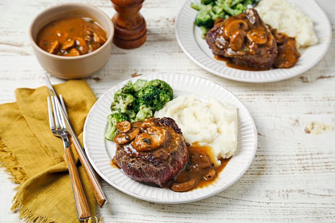 3/4th shot of Beef Tenderloin and Mushroom Sauce recipe shot on a white wooden surface; Steak served on white plates with mushrooms sauce, mashed potatoes and broccoli, along with a sliver fork and knife is placed over yellow kitchen napkin, a sprinkler and a bowl filled with dish is also visible