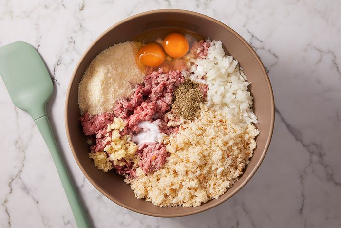 Overhead shot of a bowl sits on a marble surface filled with various ingredients and a pale green spatula rests next to it and Ingredients include ground meat bread crumbs chopped onions eggs grated cheese minced garlic and seasonings