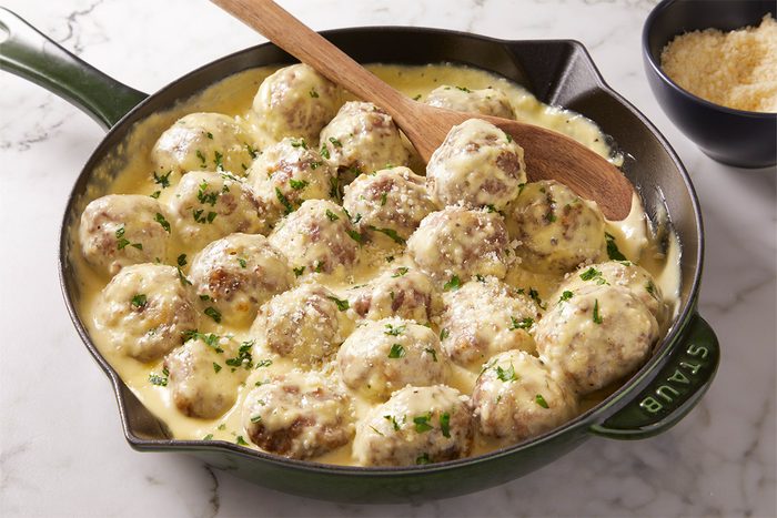 3/4th Shot of Alfredo Meatballs sit in a green skillet in creamy yellow sauce with parsley A wooden spoon is on top near a bowl of grated cheese