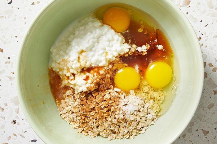 Overhead shot of a bowl containing uncooked oats, cottage cheese, two eggs, cinnamon, and liquid sweetener, all separated and ready to be mixed.