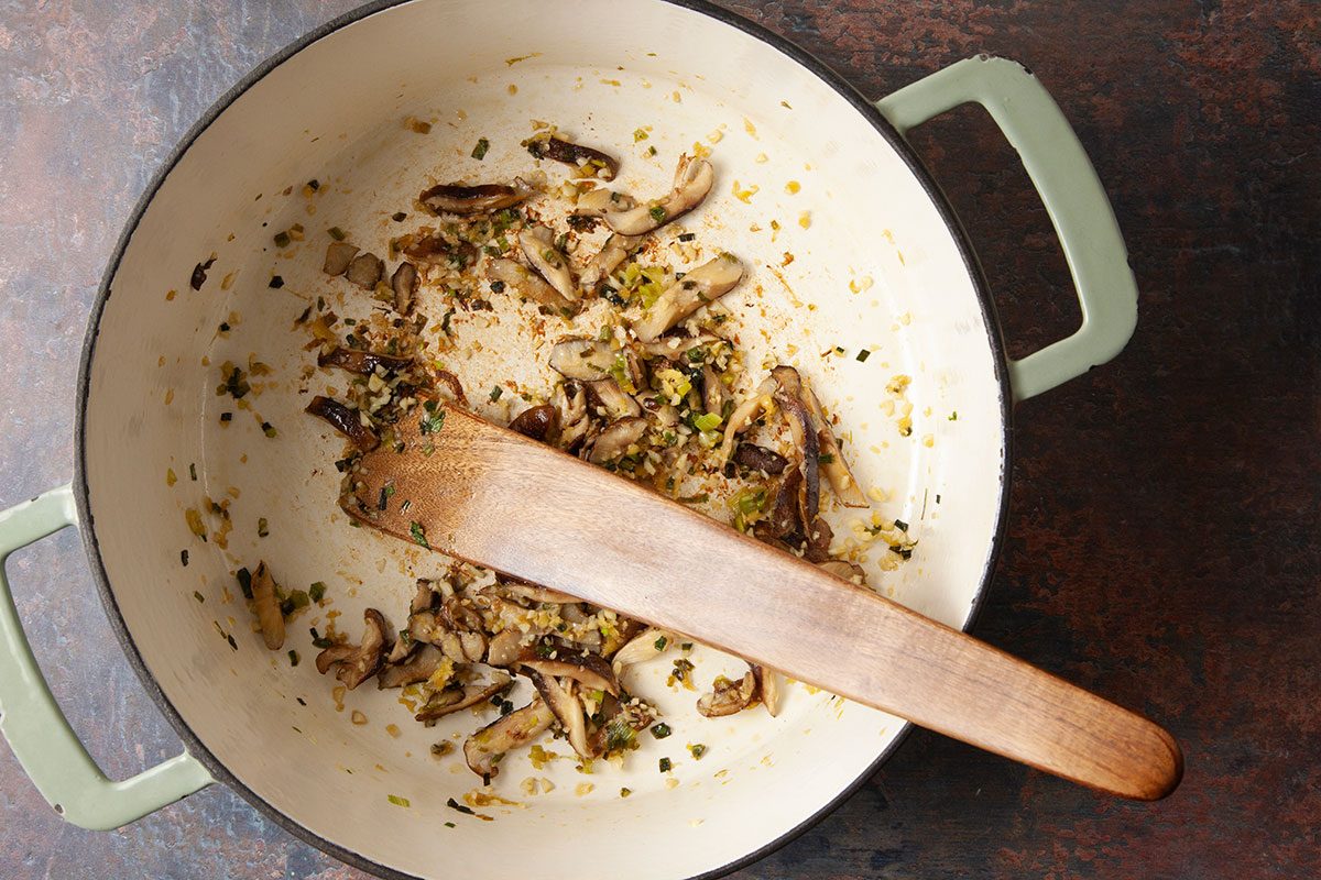 An overhead, horizontal step-by-step cooking image showing mushrooms, green onions, garlic, and fresh ginger sautéing in sesame oil inside a light-colored Dutch oven. This image captures the first stage of building flavor for Easy Dumpling Soup, highlighting the aromatic base before liquid ingredients are added.