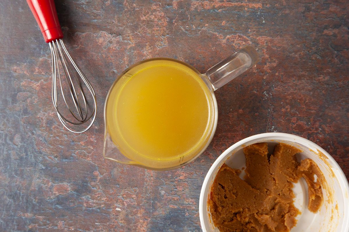 An overhead, horizontal step-by-step image featuring measured vegetable broth, miso paste, and kitchen tools arranged on a textured surface. This image highlights the prepared ingredients used to build the savory broth for Easy Dumpling Soup.