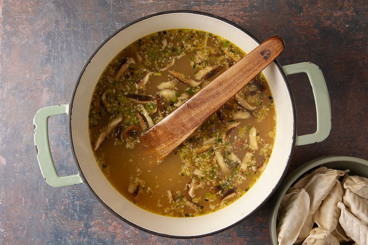 An overhead, horizontal cooking image showing vegetable broth simmering with mushrooms, green onions, garlic, ginger, and seasonings in a Dutch oven. A wooden spoon rests in the pot as the soup base for Easy Dumpling Soup develops rich flavor.