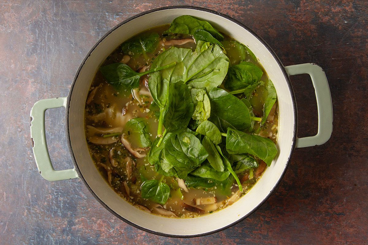 An overhead, horizontal step-by-step image showing fresh spinach added to a pot of simmering dumpling soup. The vibrant greens rest on top of the broth as the final vegetables are incorporated into Easy Dumpling Soup.