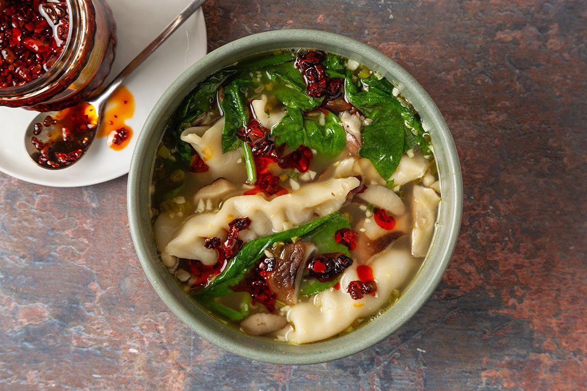 An overhead, horizontal image of finished Easy Dumpling Soup served in a bowl with tender potstickers, wilted spinach, mushrooms, and broth. Chili crisp is spooned over the top as a garnish, adding texture and heat to the comforting soup.