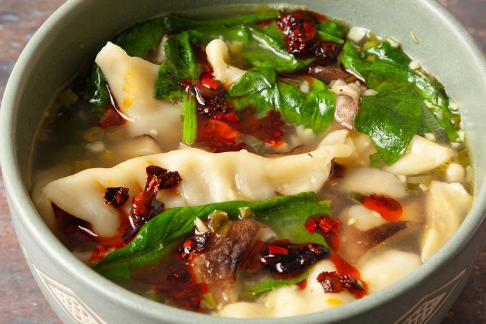 An overhead, horizontal image of finished Easy Dumpling Soup served in a bowl with tender potstickers, wilted spinach, mushrooms, and broth. Chili crisp is spooned over the top as a garnish, adding texture and heat to the comforting soup.