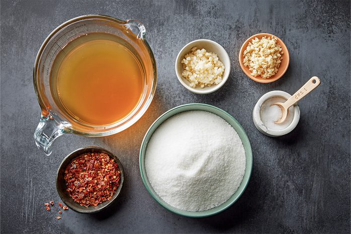 Overhead shot of a glass measuring cup with clear liquid sits beside a bowl of white sugar chopped garlic minced ginger red chili flakes and salt on a dark surface