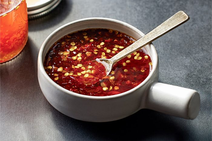 3/4th shot of SweetHot Asian Dipping Sauce fills a white bowl with a handle red chili oil and flakes swirl as a silver spoon rests inside nearby lies a glass jar with more sauce all on a dark surface