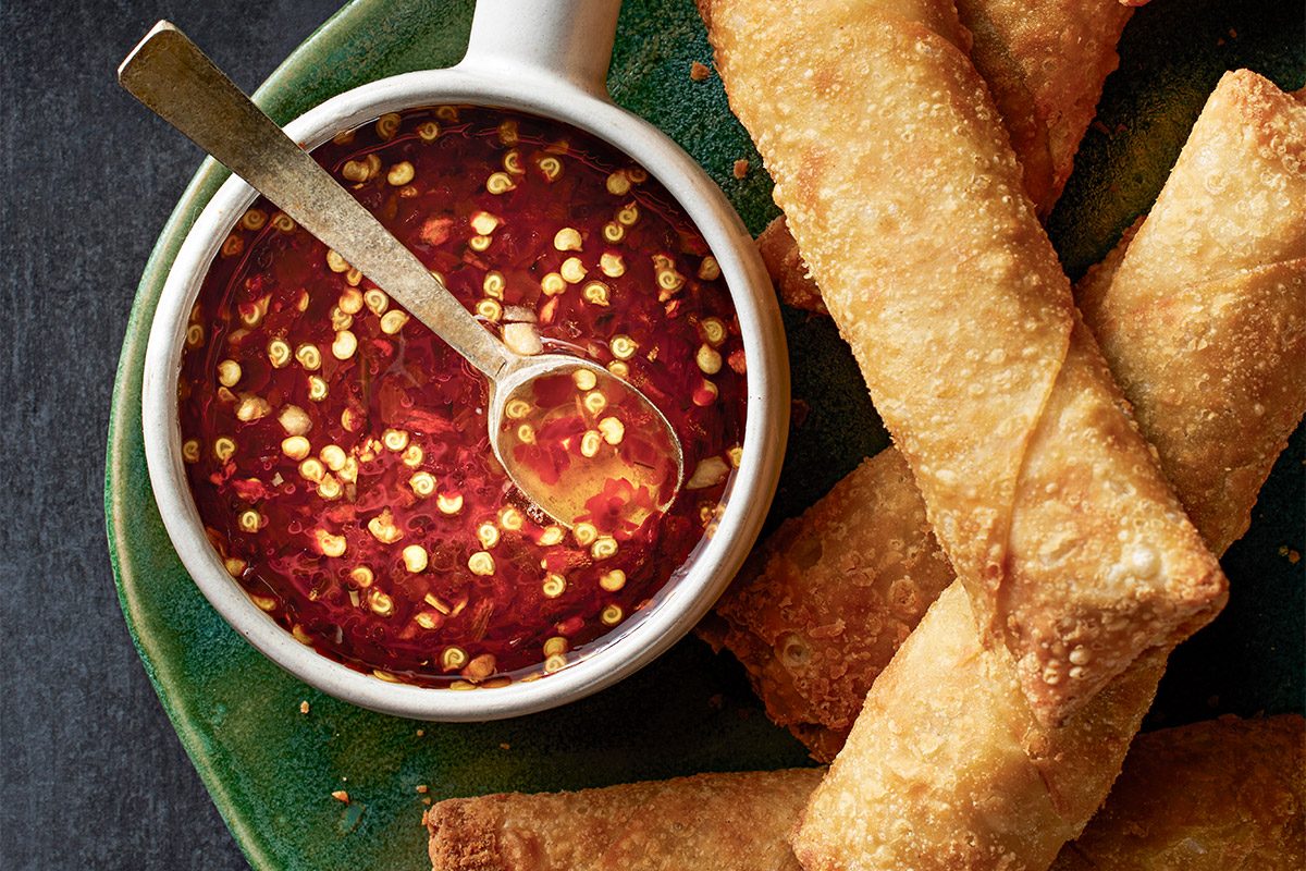 Vertical shot of a green plate golden brown egg rolls sit beside Sweet Hot Asian Dipping Sauce with a spoon nearby a cut egg roll on a black plate