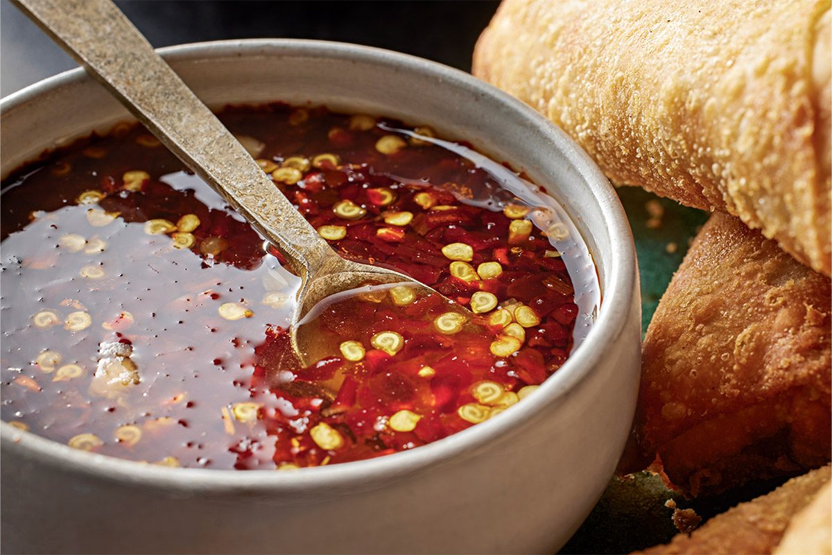 Close-up shot of a bowl of SweetHot Asian Dipping Sauce with sliced red chilies and seeds a spoon inside beside crispy fried spring rolls on green background