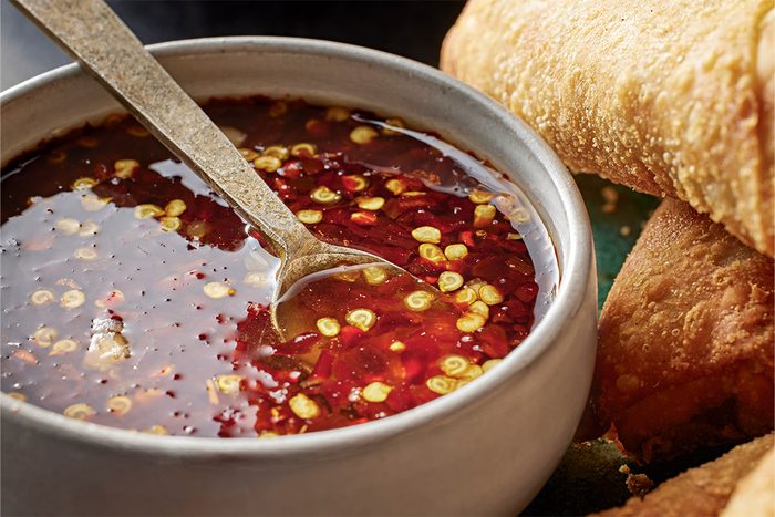 Close-up shot of a bowl of SweetHot Asian Dipping Sauce with sliced red chilies and seeds a spoon inside beside crispy fried spring rolls on green background