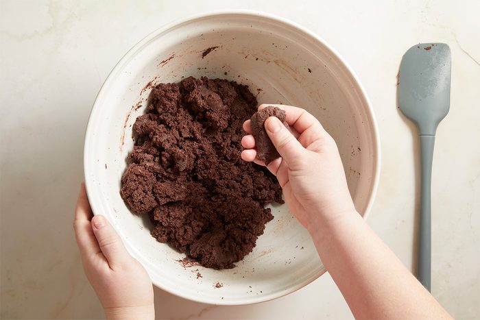 Overhead view of hands kneading and combining chocolate cake crumbs and frosting in a bowl to create a smooth cake pop dough for football-shaped cake pops.