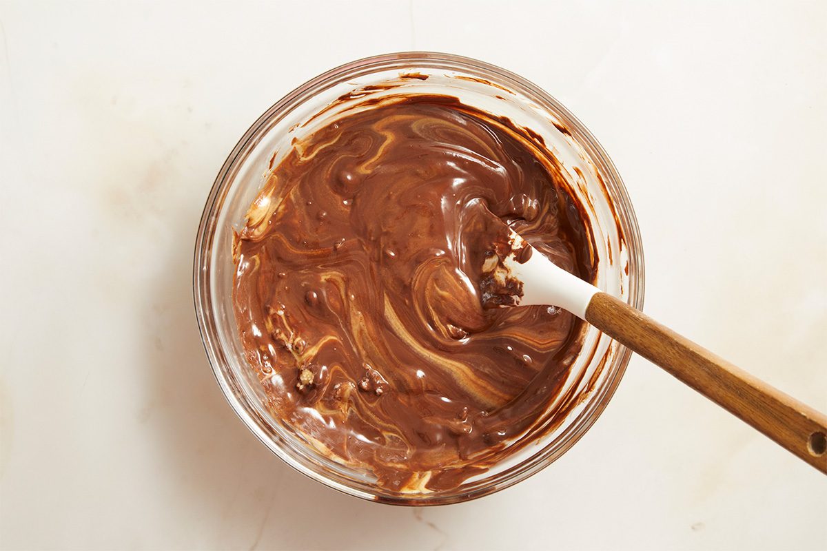 Overhead food photography of melted chocolate coating being stirred in a glass bowl, prepared for dipping football cake pops during dessert assembly.