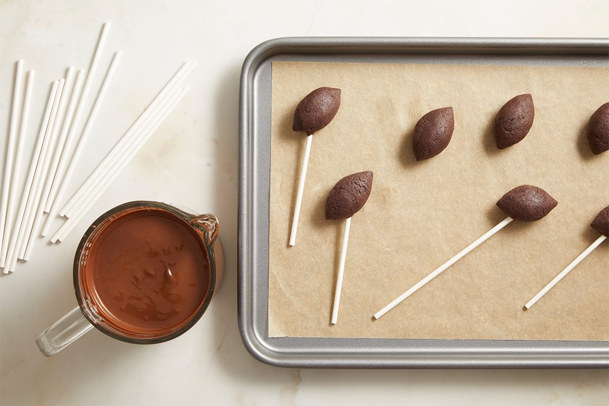 Overhead view of football-shaped cake pops with lollipop sticks inserted, arranged on parchment paper next to melted chocolate coating for dipping.
