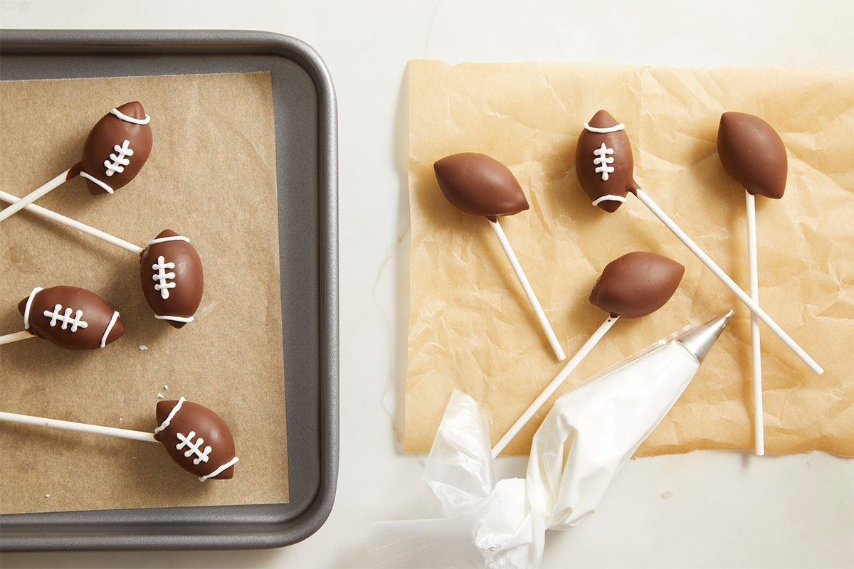 Overhead food photography of chocolate-coated football cake pops being decorated with white icing laces on parchment paper.