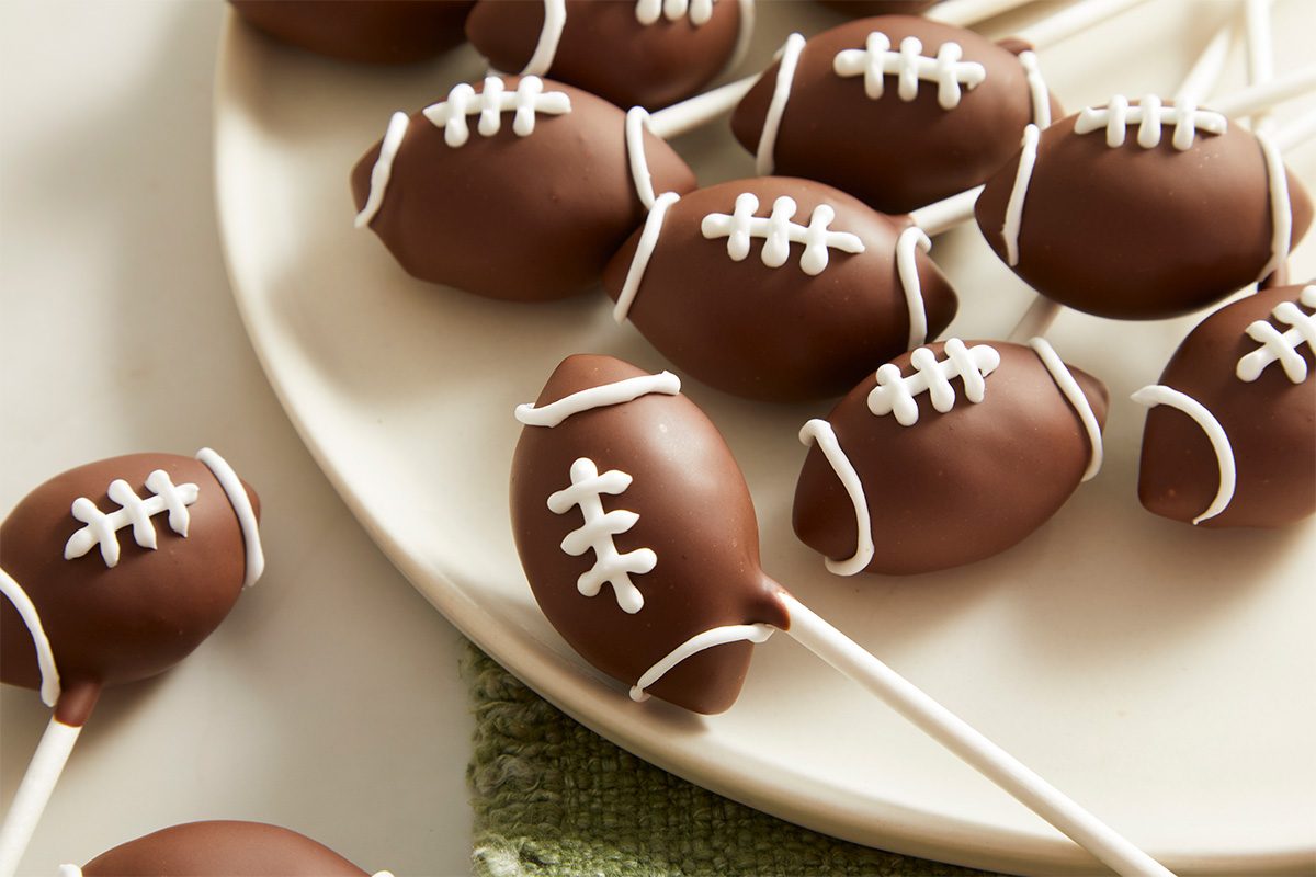 Overhead close-up food photography highlighting chocolate football cake pops with piped white icing laces displayed on a plate.