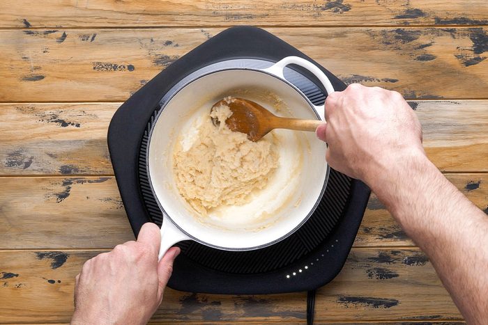 Overhead shot of a person stirs a light brown roux in a white pot using a wooden spoon over a portable stove on a rustic wood surface as they cook