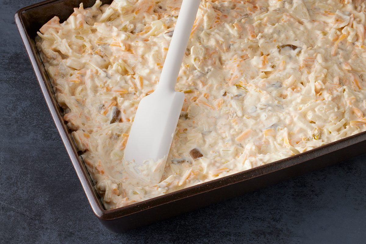 A white spatula spreads a creamy mixture evenly in a rectangular baking dish on a dark countertop.