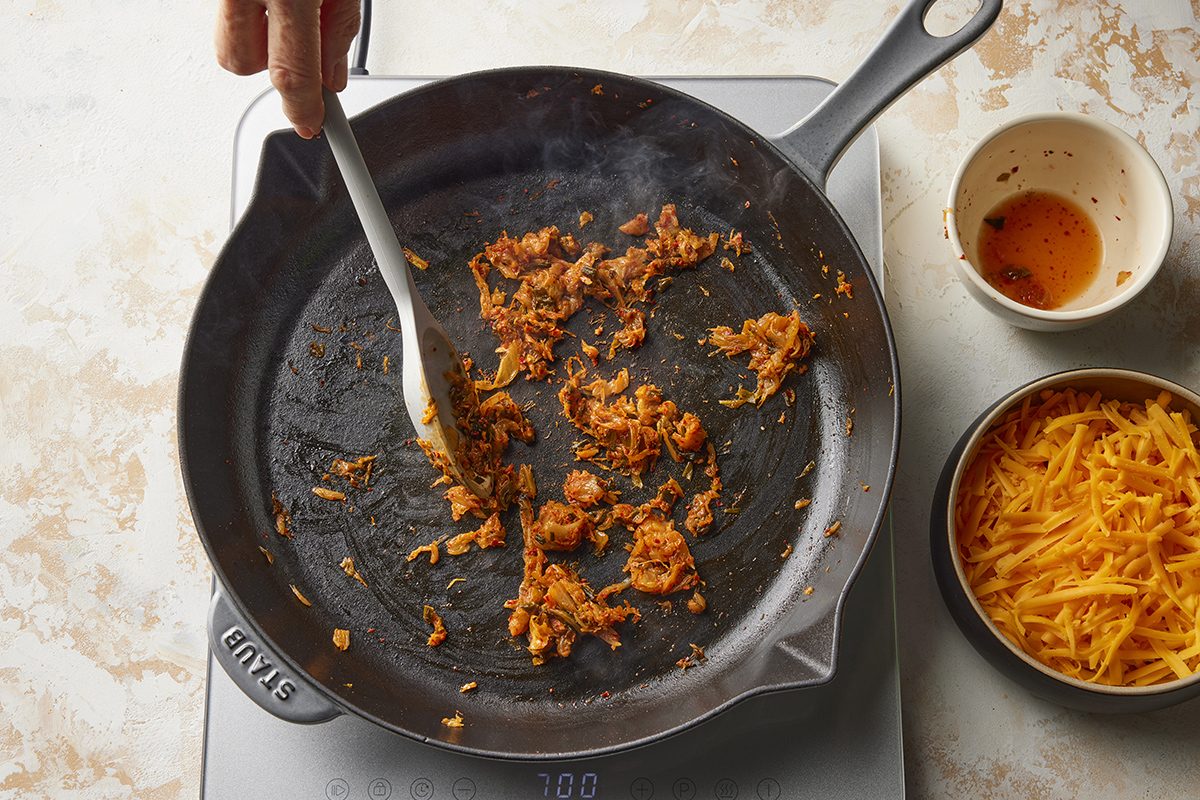 A hand stirs seasoned, cooked shredded meat in a black skillet on a stovetop. Nearby are a bowl of shredded cheddar cheese and a small bowl with leftover sauce.