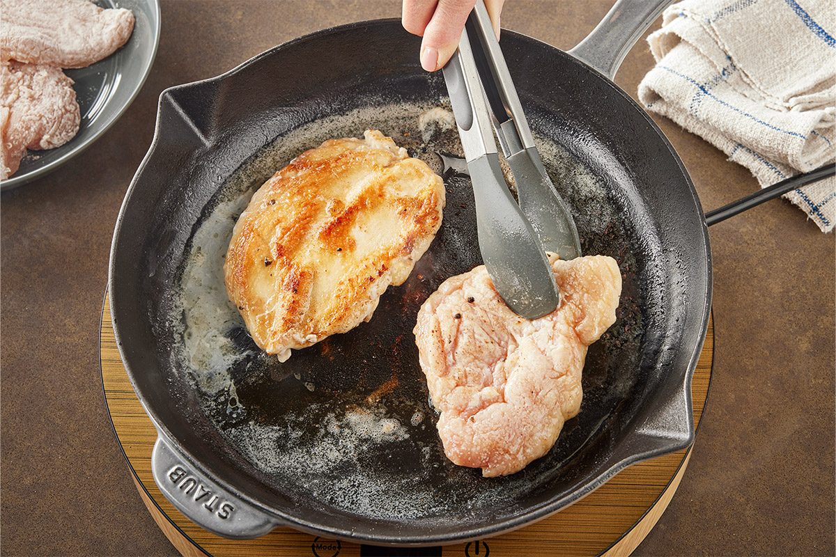 Overhead shot of a hand uses tongs to flip chicken breast in a black cast iron skillet as another piece is seared nearby a plate with raw chicken and a towel;