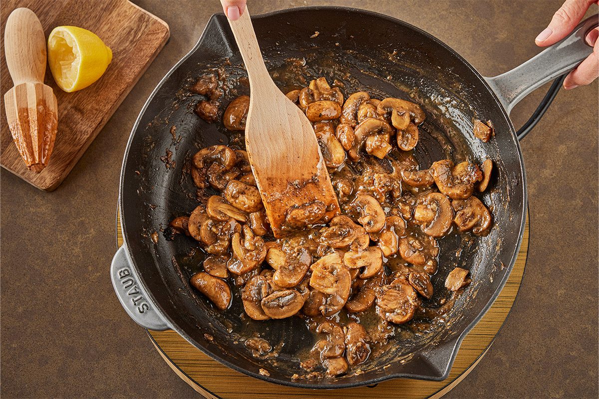 Overhead shot of a person stirs mushrooms in a skillet using a wooden spatula; A wooden citrus reamer and cut lemon are on a cutting board nearby;