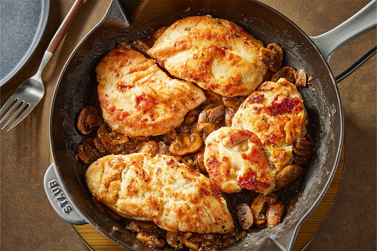 Overhead shot of Four golden-brown chicken breasts cook in a cast iron skillet with mushrooms in savory sauce; The pan sits on wood near a plate and fork;