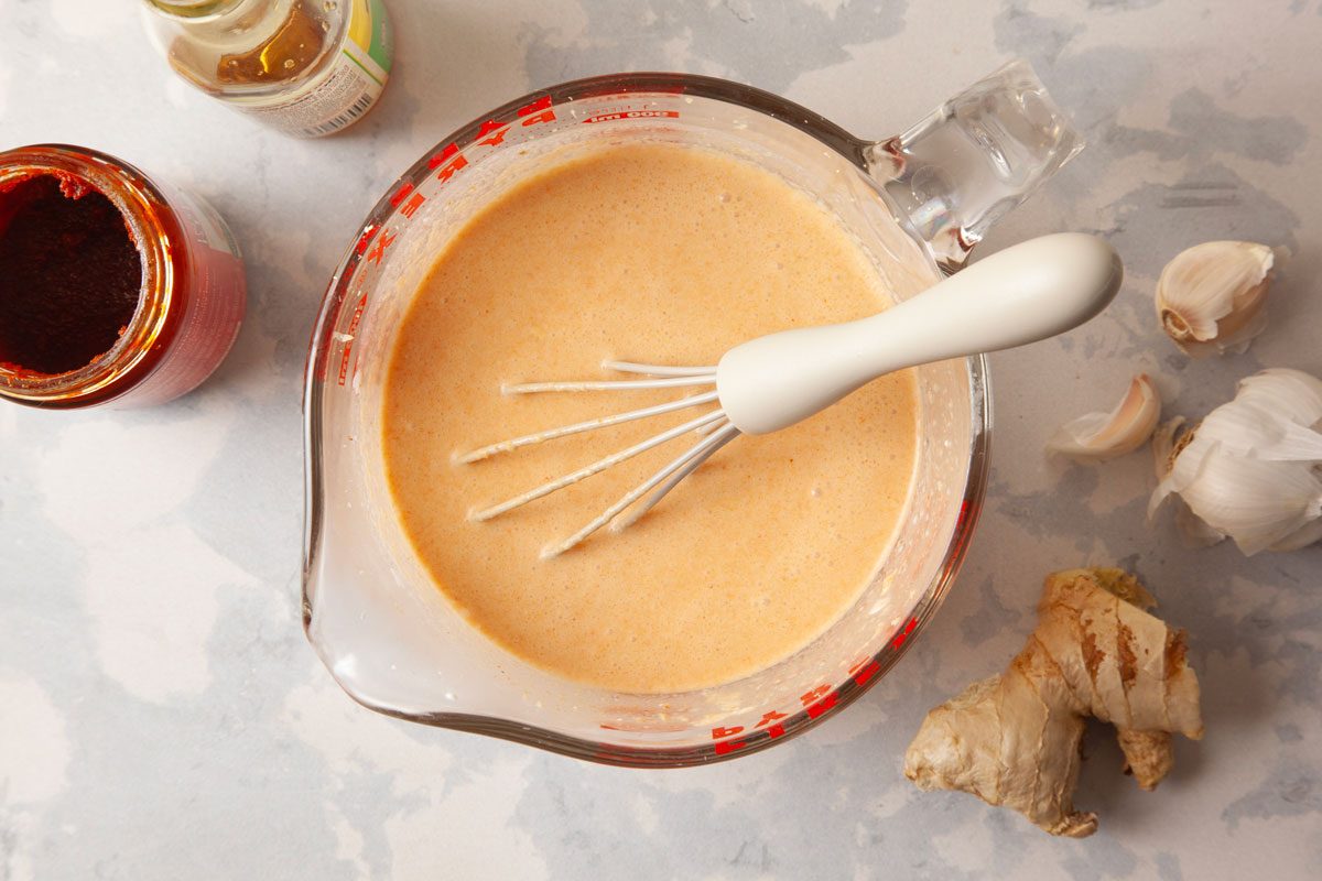 Overhead shot of a metal whisk sits in a clear glass measuring cup with smooth orange mixture surrounded by garlic cloves ginger an open red paste jar and a bottle on a pale countertop