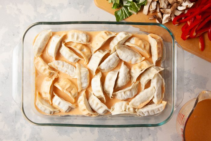 Overhead Shot of a glass dish holds dumplings in orange sauce nearby a cutting board with green vegetables mushrooms red peppers and a cup of sauce