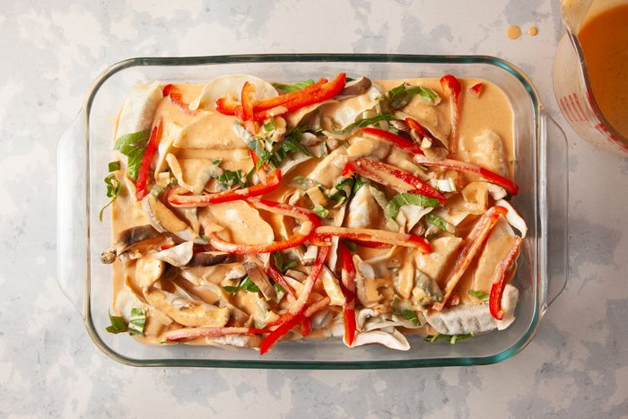 Overhead shot of a transparent glass baking dish contains layers of uncooked enchiladas orange sauce sliced red peppers green herbs rests on a pale countertop