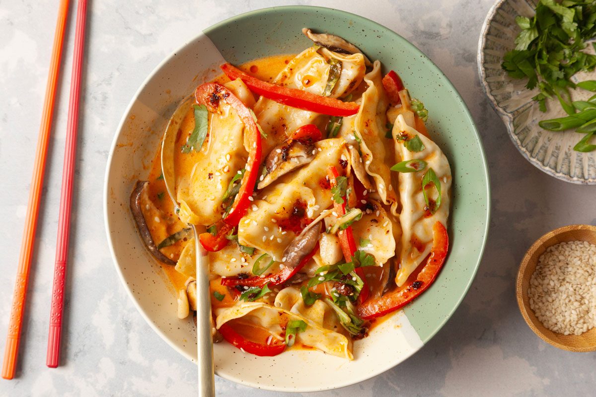 Overhead shot of a One Pan Dumpling Dinner shows dumplings in creamy orange sauce with red peppers mushrooms green onions sesame seeds chopsticks herbs nearby