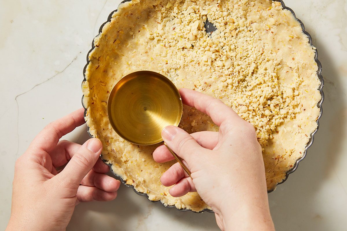 Overhead, horizontal step-by-step image showing hands pressing a crumb mixture of flour, toasted sliced almonds, butter, sugar, and orange zest into a tart pan to form the crust for a passion fruit tart.