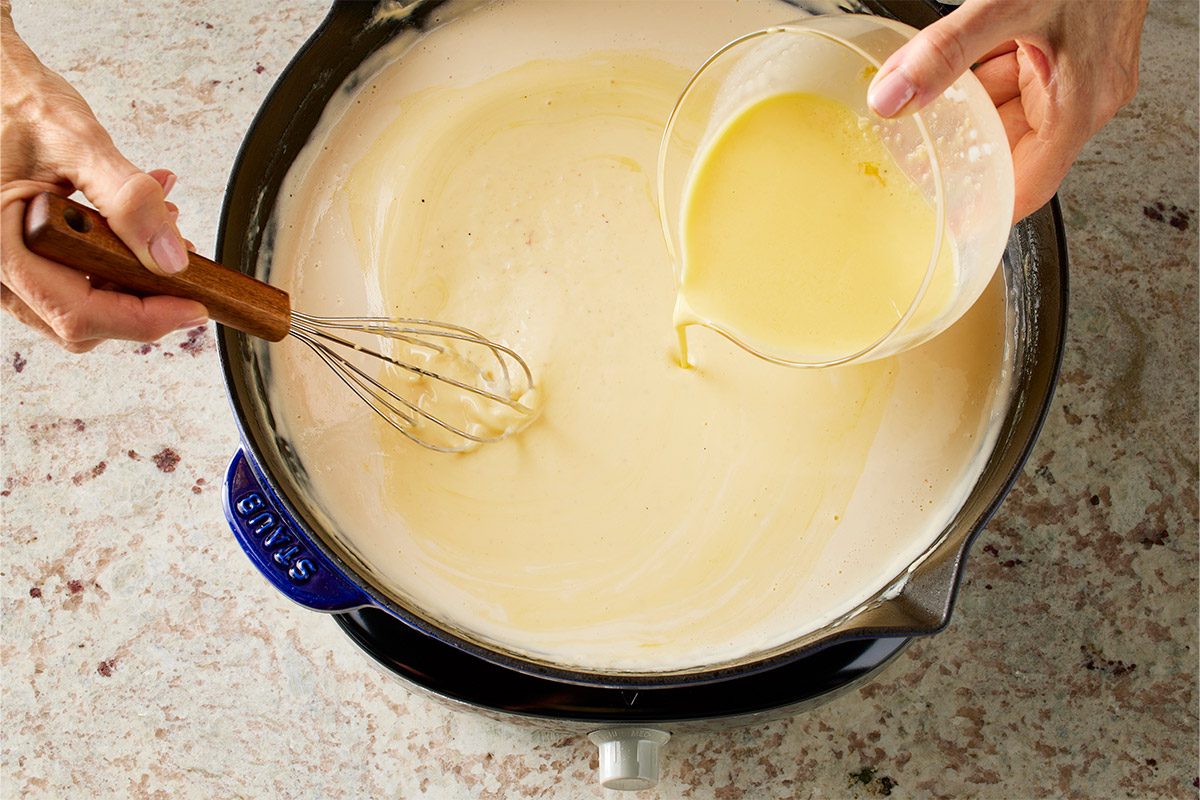 Overhead shot of a person stands over a grey marble countertop whisking a pale creamy mixture in a silver saucepan while pouring liquid from a white bowl