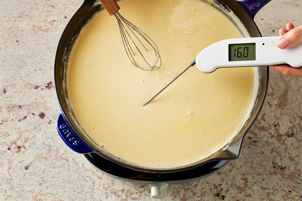 Overhead shot of a human hand grips a digital thermometer displaying 160 degrees Fahrenheit above a silver pot of smooth yellow mixture on a stovetop while a metal whisk stirs the creamy contents the stove surface appears dark and sleek with minimal background objects present in the clear kitchen setting