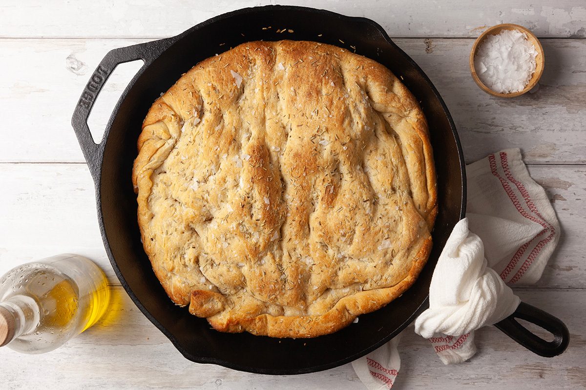 Overhead shot of shortcut pizza dough focaccia, golden-brown and baked in a cast iron skillet, set on a light wooden surface with a bottle of olive oil, a bowl of salt, and a kitchen towel nearby;