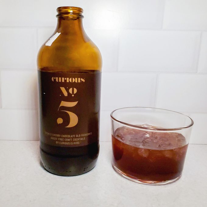 A brown glass bottle labeled "Curious No. 5" is next to a clear glass containing a dark brown beverage with ice, set on a white countertop with a white tiled wall in the background.
