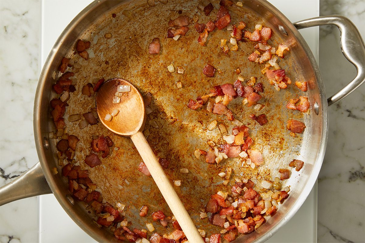 Overhead view of chopped thick-sliced bacon, onion, garlic, and crushed red pepper flakes sautéing in a wide skillet as the base for a creamy vodka sauce. The rendered bacon fat coats the pan while aromatics soften and caramelize, marking the first step in preparing Vodka Sauce with Cottage Cheese.