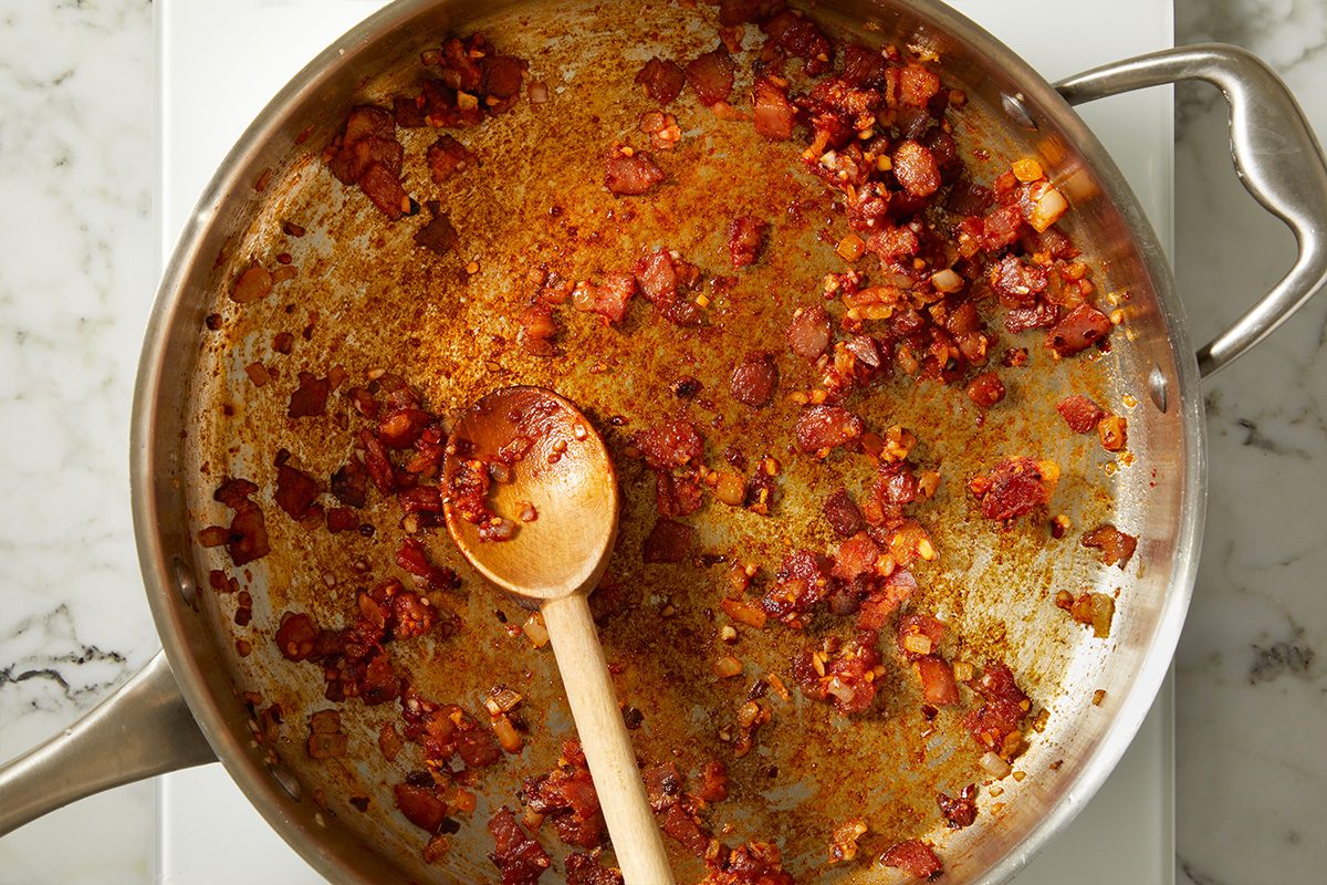 Top-down view of sautéed bacon, onions, and garlic continuing to cook in a skillet, deepening in color and flavor. The mixture shows increased browning and texture as the aromatic base develops for a rich vodka pasta sauce.