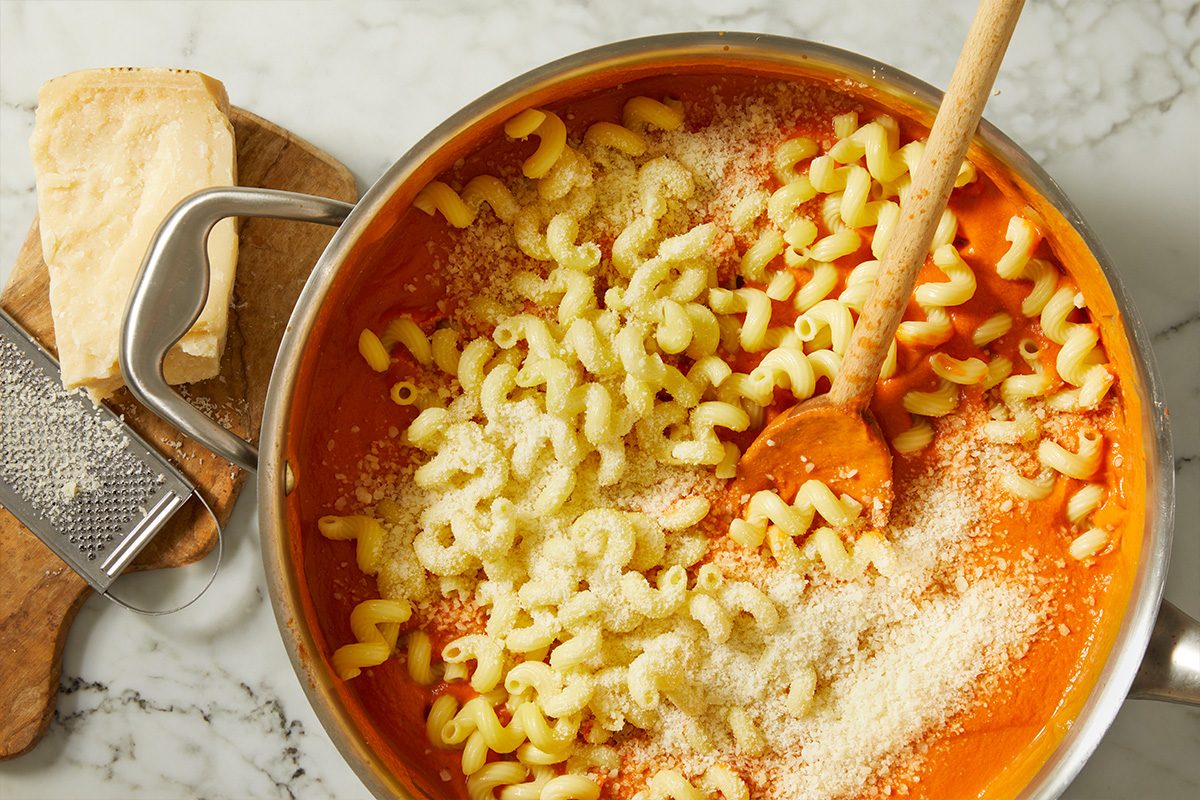 Overhead shot of cooked pasta added directly to the vodka sauce, with grated Parmesan cheese sprinkled on top. The pasta is tossed to evenly coat each piece in the creamy tomato sauce.
