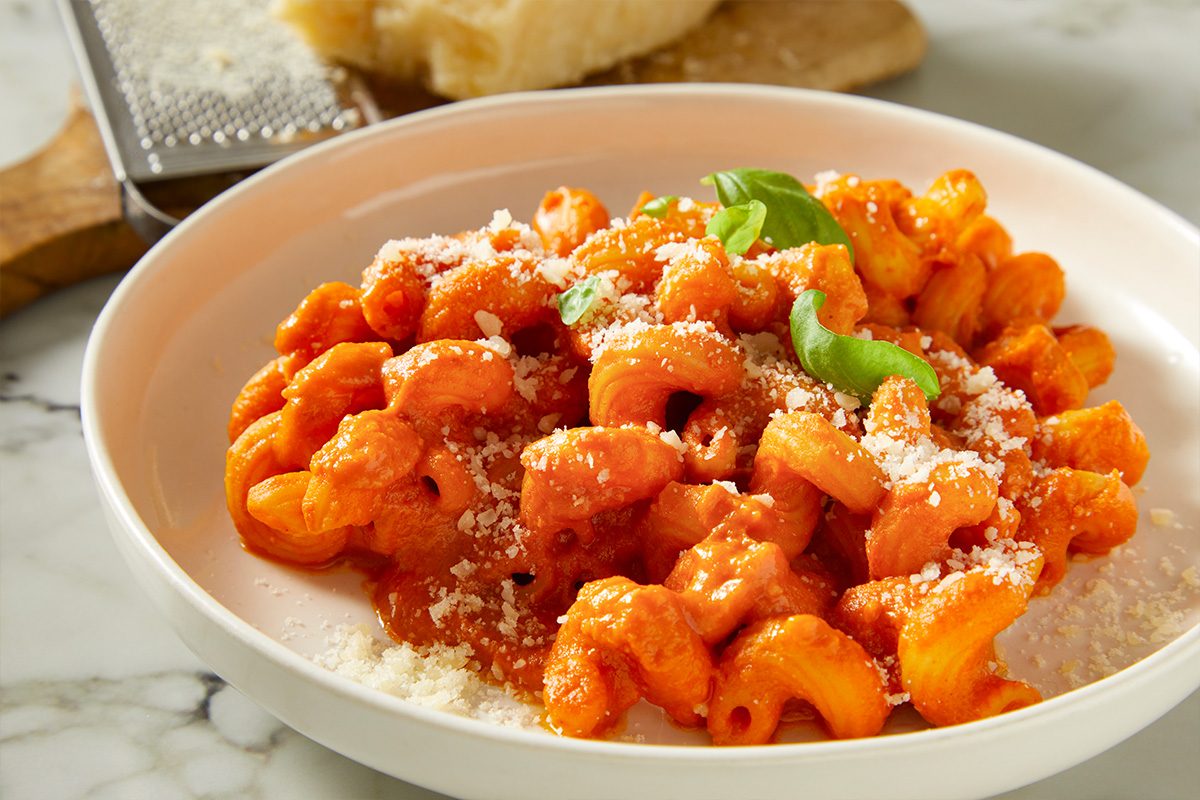 Close-up of a bowl filled with creamy vodka pasta, topped with grated Parmesan and fresh herbs. The sauce appears rich and smooth with visible bacon pieces throughout.