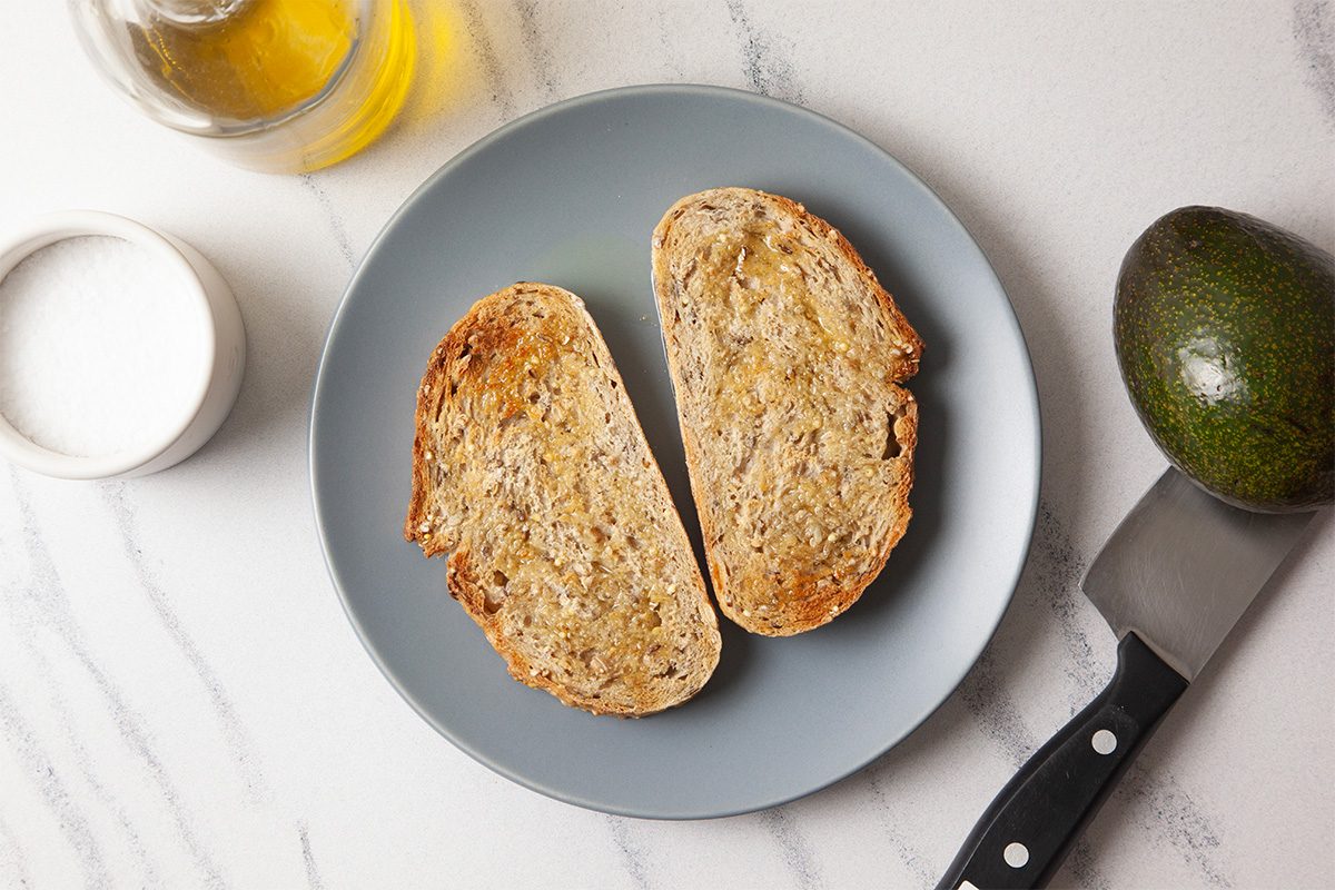 Overhead horizontal image of two slices of hearty toasted bread arranged on a neutral plate atop a light marble surface. The scene is minimal and clean, establishing the base ingredient for a simple avocado toast preparation.
