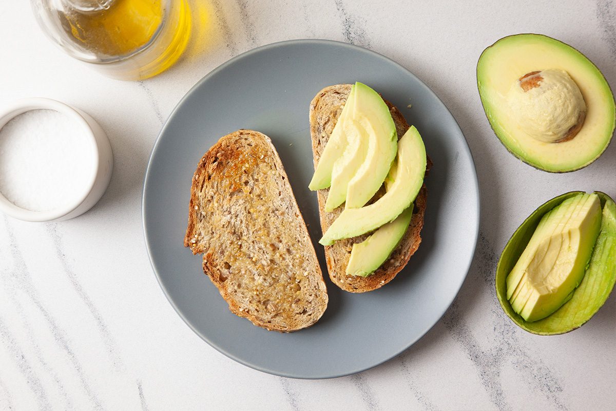 Overhead horizontal image showing toasted bread topped with freshly sliced ripe avocado. Whole and halved avocados are styled nearby, emphasizing freshness and preparation for avocado toast assembly.