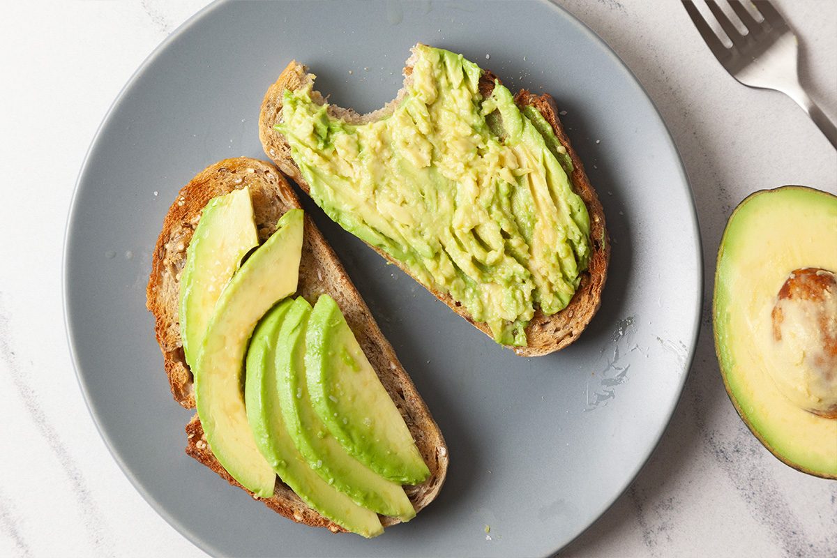 Overhead horizontal image of avocado toast with mashed avocado spread generously across toasted bread, paired with sliced avocado on the side to show texture variation.