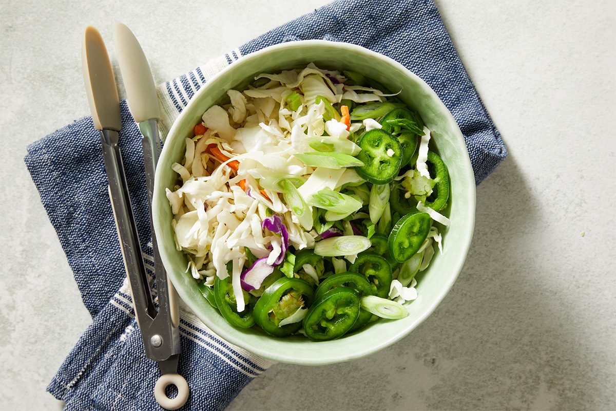 An overhead, horizontal step-by-step image showing a bowl of coleslaw mix combined with sliced green onions and jalapeño peppers. This fresh slaw is prepared as a topping for Spicy Corned Beef Tacos.