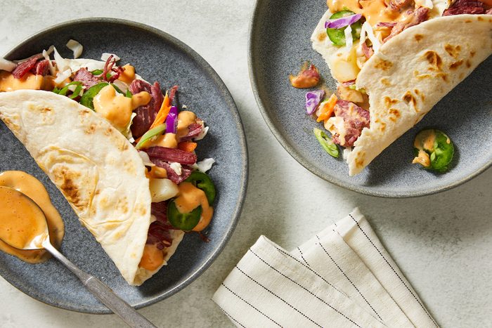 An overhead, horizontal image featuring two plated Spicy Corned Beef Tacos served with extra spicy Thousand Island sauce on the side. The tacos are filled with corned beef, potatoes, and fresh slaw.