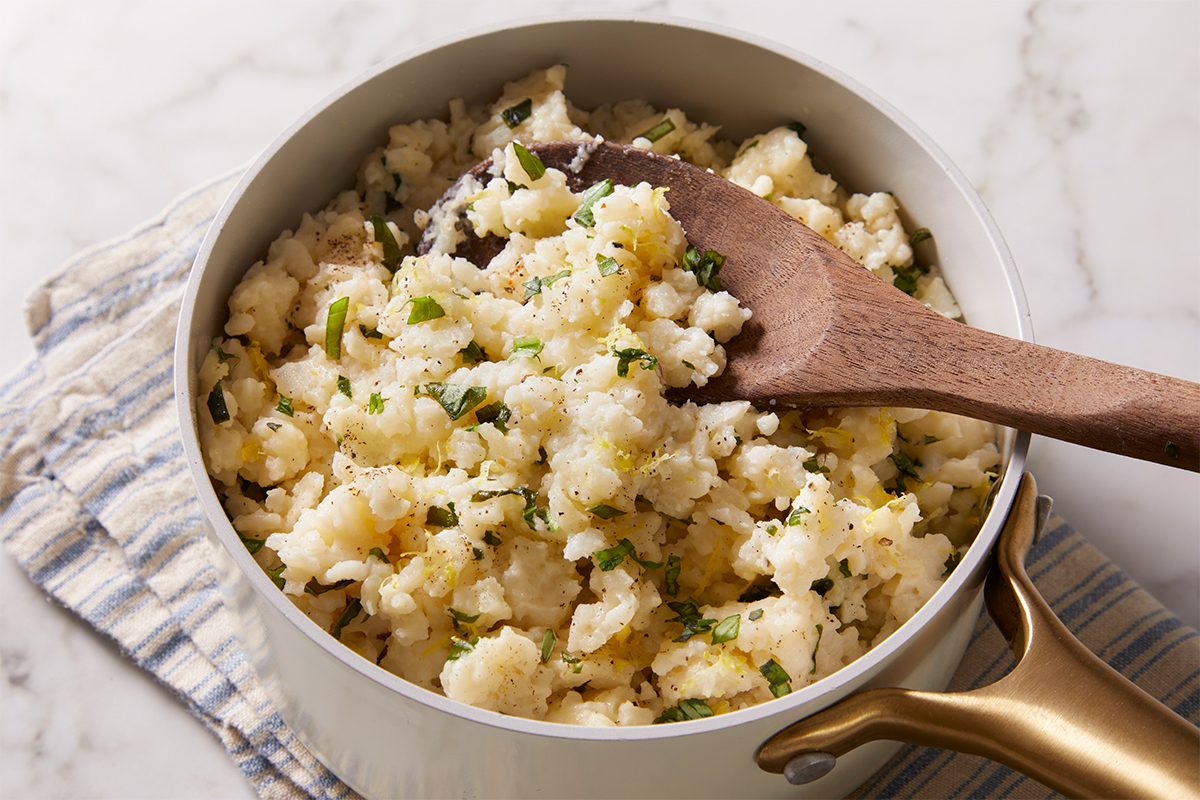 3/4 shot of a pot of Creamy Lemon Rice being stirred with a wooden spoon, showing fluffy texture and light herbs on top.