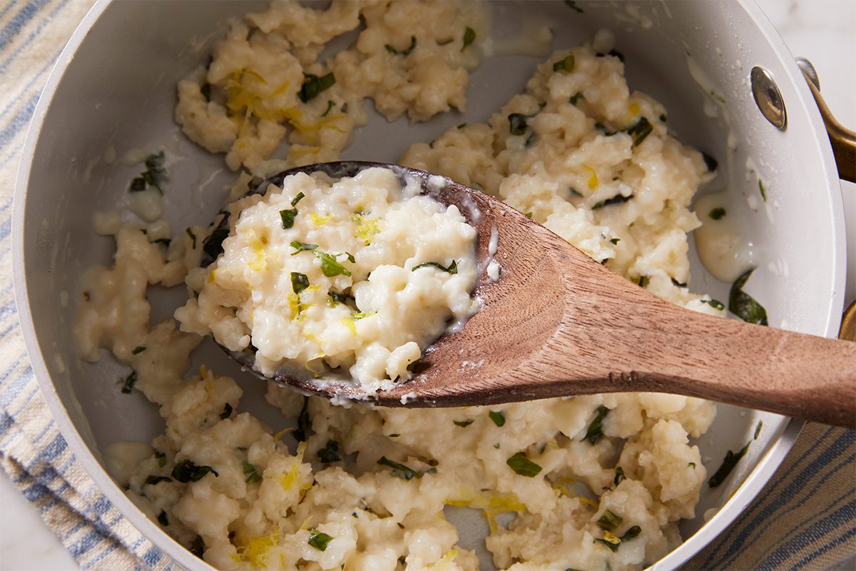 Close-up overhead shot of Creamy Lemon Rice in a saucepan, with a wooden spoon scooping a portion, showing creamy consistency and bits of herbs.