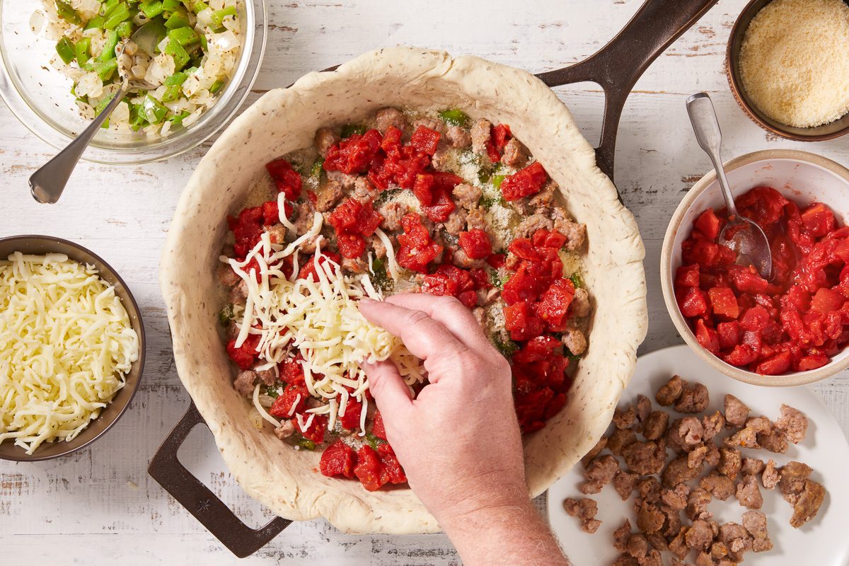 mozzarella cheese being sprinkled over layered vegetables, sausage and tomatoes